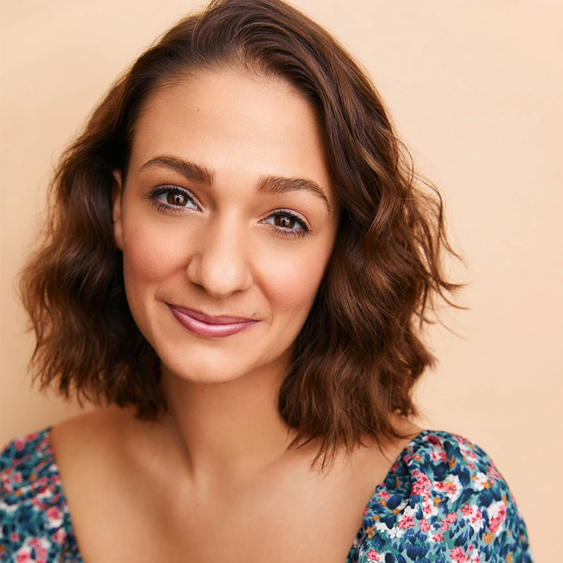 A young woman with wavy brown hair smiles gently at the camera. She has light skin, brown eyes, and is wearing a floral-patterned top. The background is a soft peach color, creating a warm and inviting atmosphere.