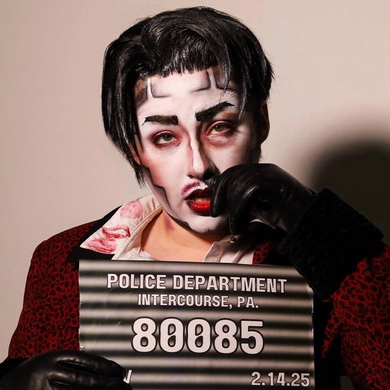 A person in dramatic, stylized makeup and a red leopard-print jacket poses biting a black-gloved finger. They hold a mugshot sign reading 