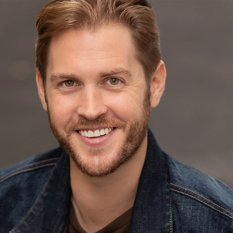 Christopher Brian Williams, a smiling man with light brown hair and a beard, wearing a dark denim jacket over a brown shirt, poses in front of a softly blurred gray background. He looks directly at the camera, exuding a friendly, approachable demeanor.