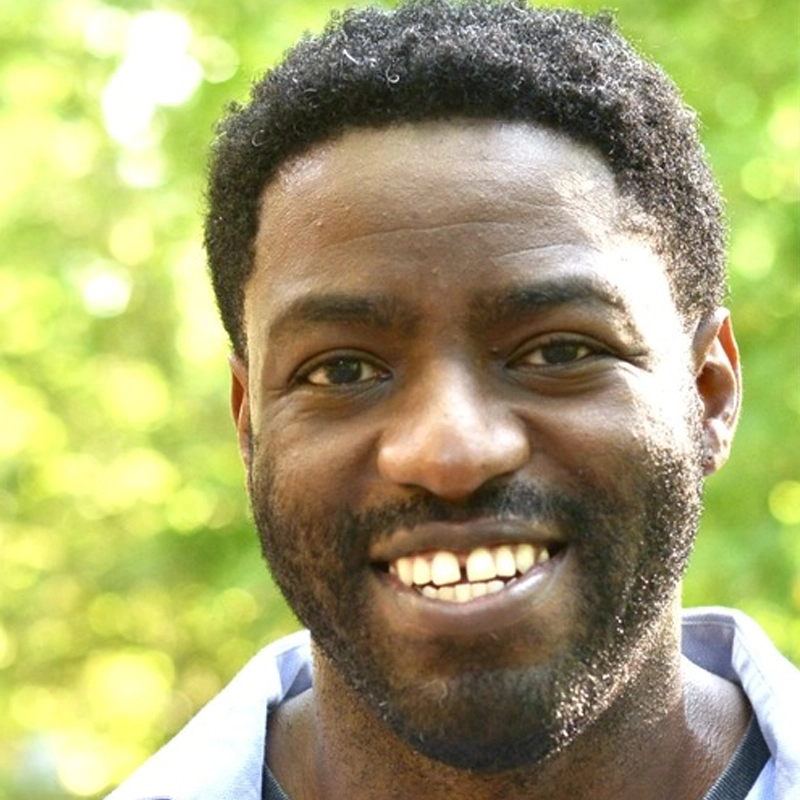 Chukwuma Obasi, a man with short curly hair and a beard, smiles at the camera outdoors. He is wearing a light blue collared shirt, surrounded by green foliage brightly lit by sunlight.