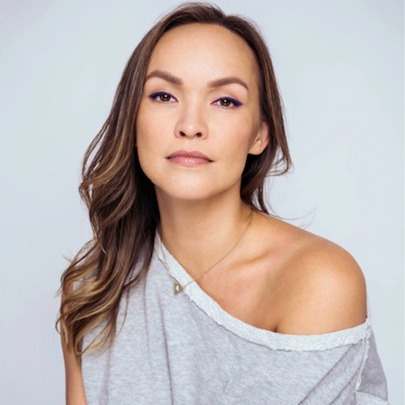 A woman with long brown hair and light makeup gazes calmly at the camera. Nicci Claspell wears an off-the-shoulder gray top and a delicate pendant necklace, set against a plain light gray background. Her expression is neutral and composed.