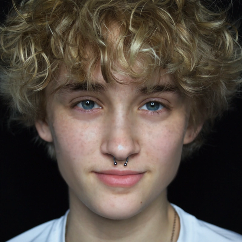 A young person with curly blond hair and light skin looks straight at the camera, smiling slightly. They have blue eyes, faint freckles, and wear a silver septum ring. They are dressed in a white shirt against a solid black background.