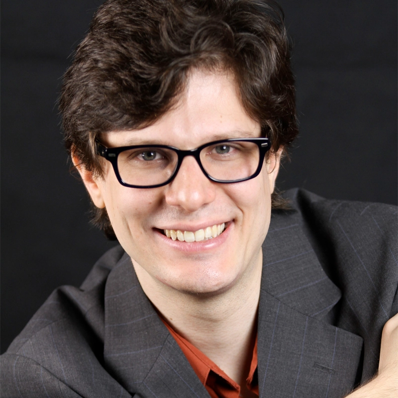 A smiling person with light skin and short, dark, wavy hair wears black-rimmed glasses, a grey suit jacket, and an orange shirt. Deniz Cordell is posed against a solid black background, appearing friendly and approachable.