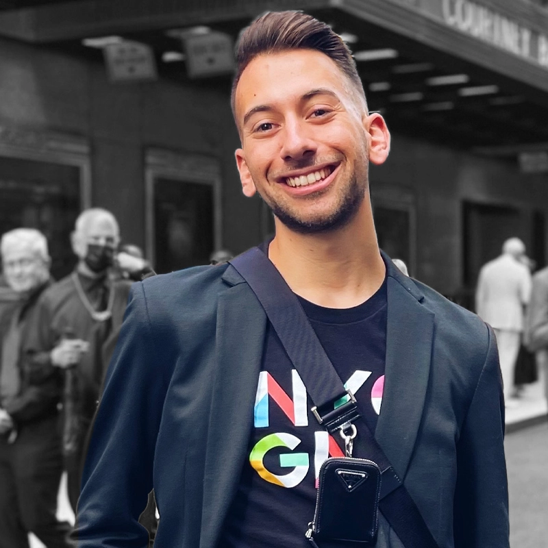 A smiling young man with short hair stands outdoors wearing a dark blazer over a colorful 