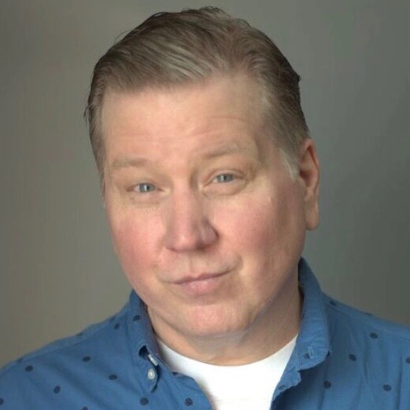Ryan Williams, a middle-aged man with light skin and short, neatly-combed blond hair, looks at the camera. He wears a blue button-up shirt over a white T-shirt and has a slight smirk on his face against a neutral gray background.