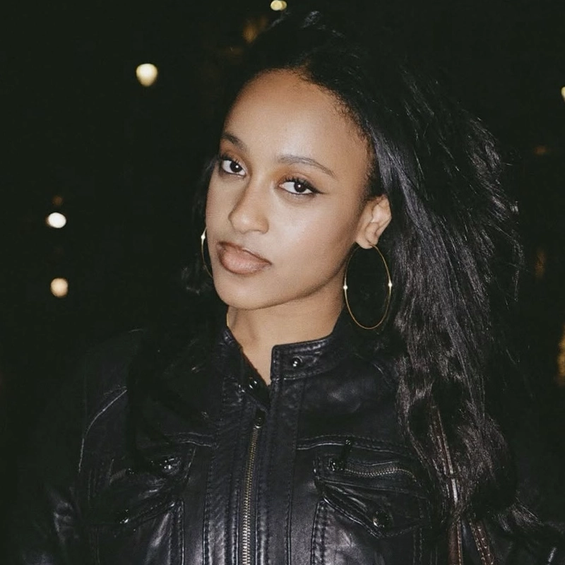 A young woman with long black hair and gold hoop earrings wears a black leather jacket. She stands outdoors at night, looking at the camera with a slight smile and neutral expression. Blurred lights are visible in the dark background.