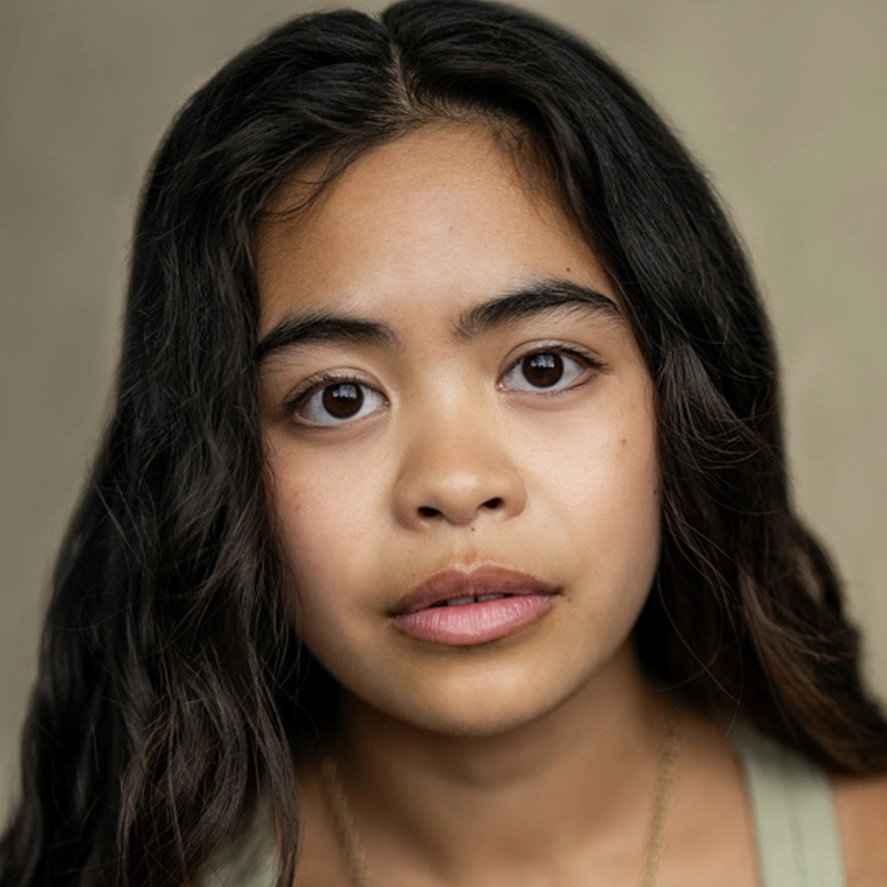 A young woman, Jennifer Duka, with long, wavy dark hair and smooth skin looks directly at the camera with a neutral expression. She has full eyebrows, large dark eyes, and natural lips against a softly blurred beige background.