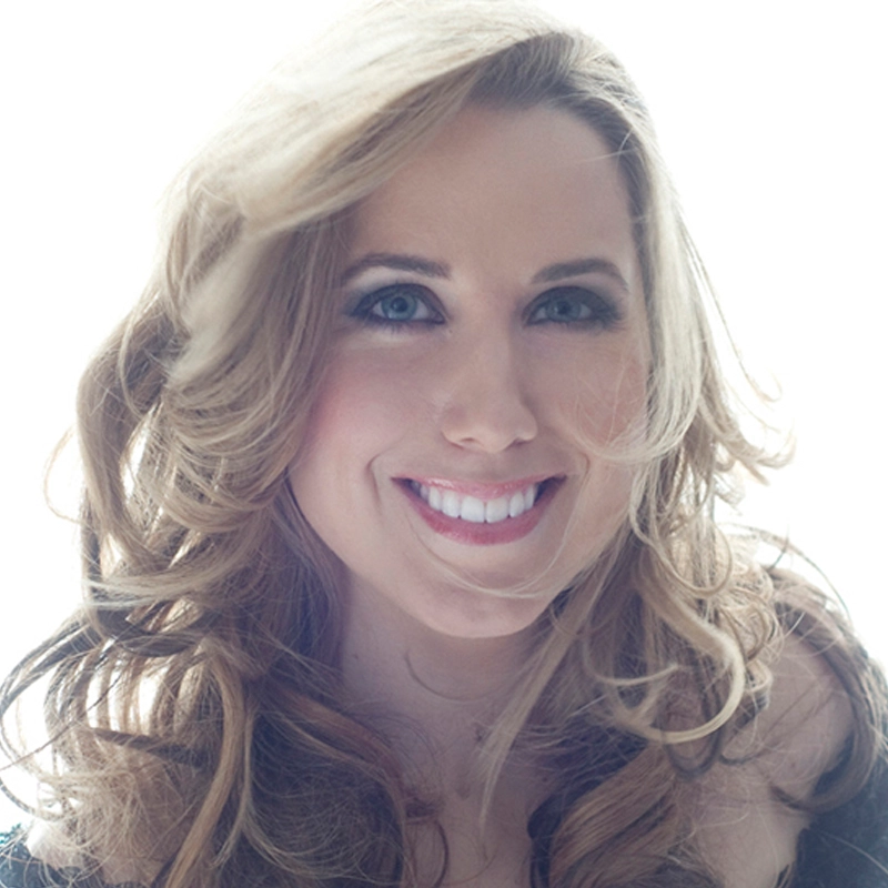 A woman with long, wavy blonde hair and blue eyes smiles warmly at the camera. Hayden DeWitt has dramatic eye makeup and is lit from behind by bright, soft light, giving the image a glowing, airy feel.