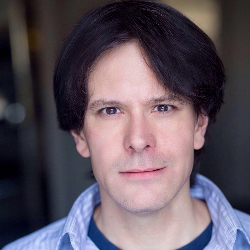 A man, identified as Ryan Williams, with pale skin and straight, dark brown hair stands indoors. He is wearing a blue, open-collar shirt over a dark t-shirt and looks directly at the camera with a neutral expression. The background is softly blurred.