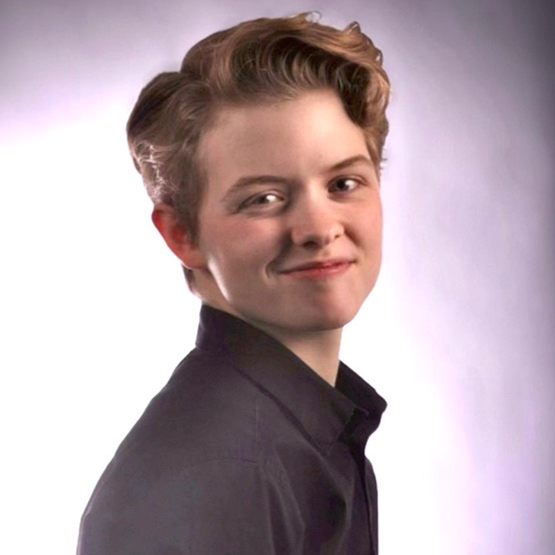 A young person with short, wavy light brown hair, fair skin, and a slight smile looks over their shoulder. They are wearing a dark collared shirt and standing against a soft, light gray background with gentle lighting.