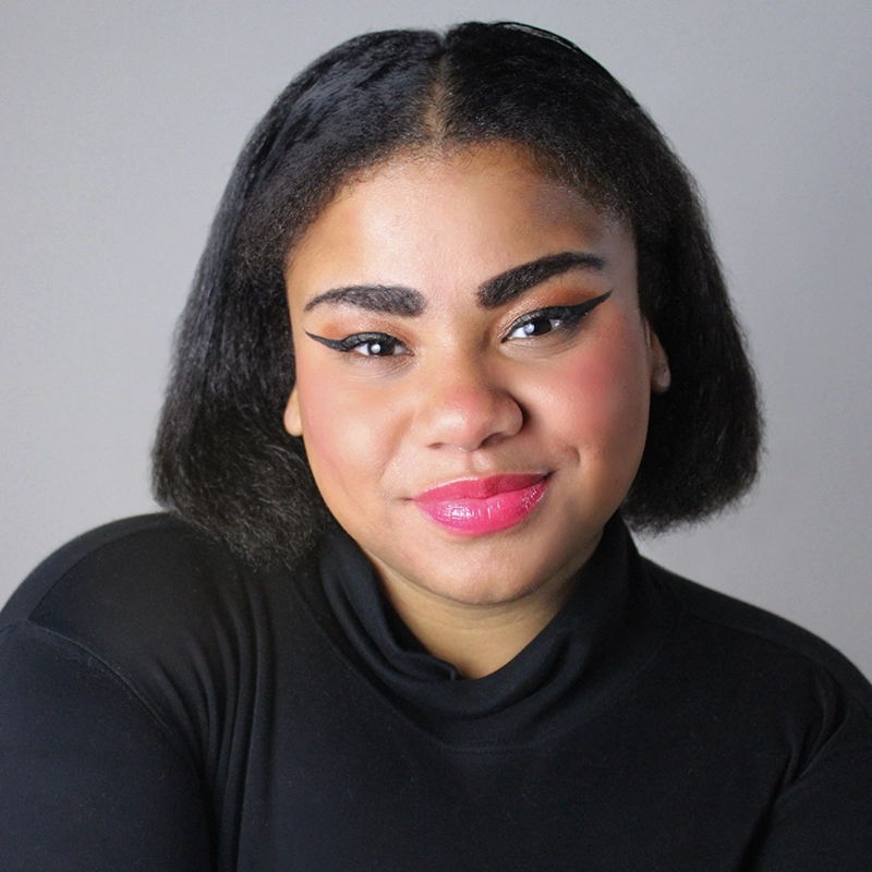 Francesca Parker, a woman with medium brown skin and short, straight black hair, smiles gently. She wears a black turtleneck, bold winged eyeliner, pink blush, and glossy pink lipstick against a plain light gray background.