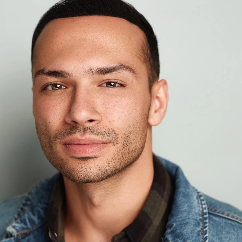A man with short dark hair and trimmed facial hair looks directly at the camera with a neutral expression. He is wearing a denim jacket over a dark plaid shirt. The background is plain and light-colored.
