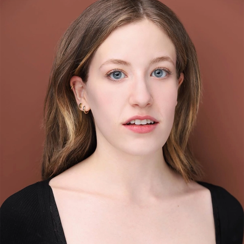 Eve Harrison, a young woman with fair skin, straight light brown hair, and blue eyes, looks at the camera with a neutral expression. She is wearing a black top and gold hoop earrings, standing in front of a plain brown background.