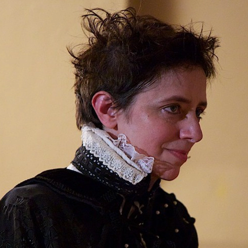 Hayden DeWitt, with short, tousled dark hair, wears an elaborate black garment and a white lace ruffled collar, looking to the side with a faint smile. Soft lighting highlights their features against a plain beige background.