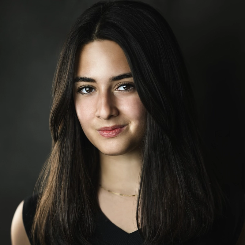 Ashley Hoberman, a young woman with long, straight dark brown hair and fair skin, poses against a dark background. She wears a sleeveless black top, a delicate gold necklace, and has a gentle, confident smile.
