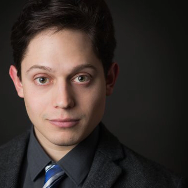 A young man with fair skin and short dark hair wears a dark grey suit, black shirt, and blue striped tie, looking directly at the camera with a slight smile against a dark, plain background.
