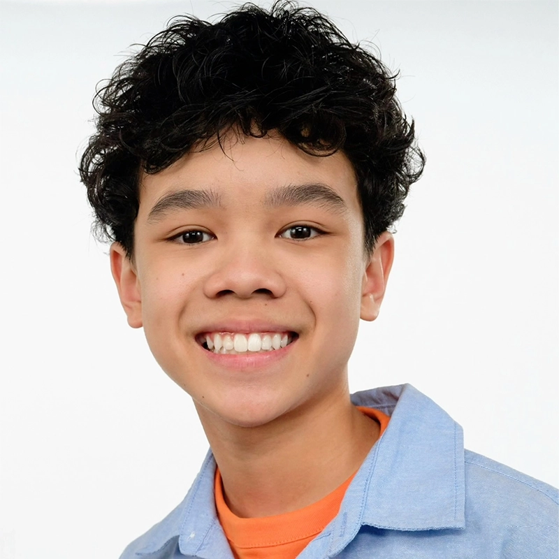 A smiling teenage boy with curly dark hair, brown eyes, and light skin wears a light blue collared shirt over an orange T-shirt. The background is plain white, emphasizing his friendly expression and casual appearance.