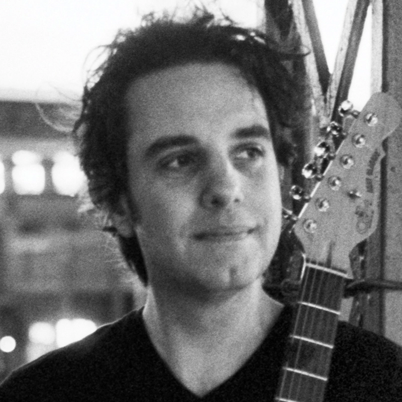 A young man with wavy dark hair and a slight smile, Ryan Williams, holds an electric guitar upright. He wears a dark shirt. The background is out of focus, showing a structure with beams and soft, glowing lights in black and white.