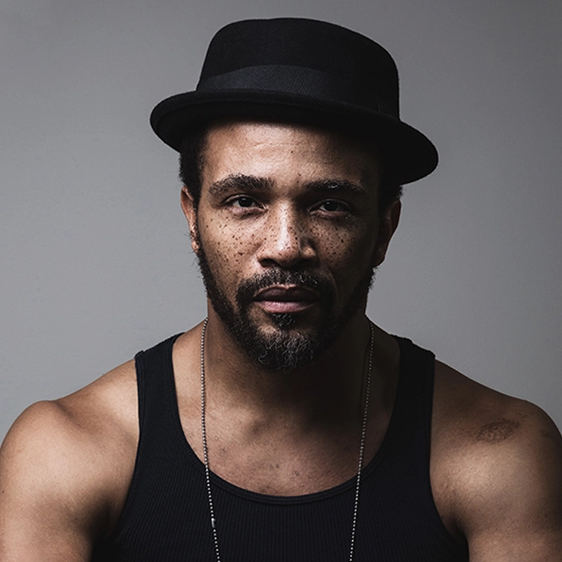 Hayden DeWitt, with a short beard and mustache, wears a black tank top and a black fedora hat. He has freckles, short curly hair, and maintains a neutral expression while looking directly at the camera against a plain gray background.