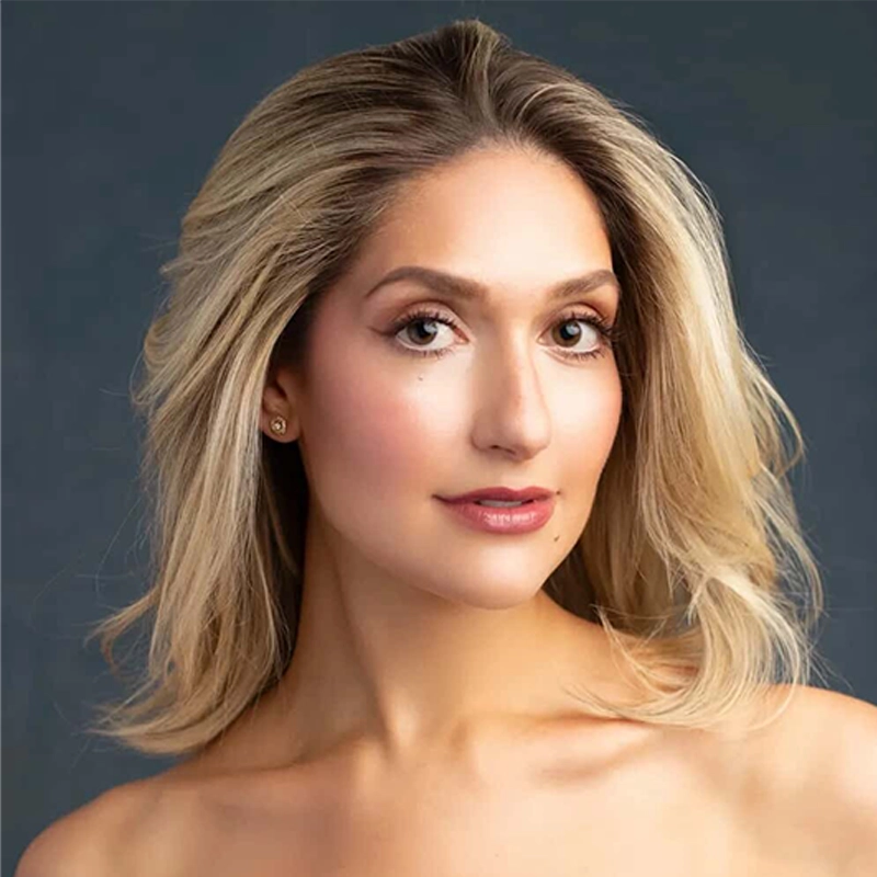 A woman with shoulder-length, wavy blonde hair and glowing skin looks towards the camera. Jamie Glickman has light makeup, brown eyes, groomed brows, and wears small stud earrings. The dark blue-gray background frames her bare shoulders.