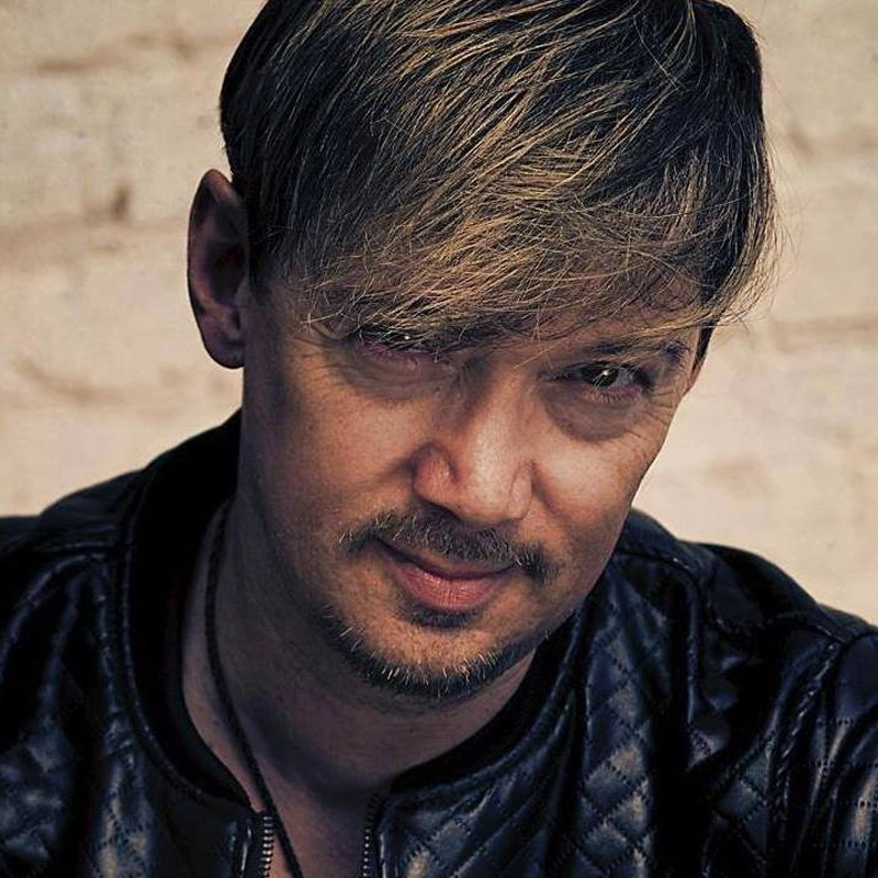 Hayden DeWitt, with light brown hair and a short beard, looks into the camera. He wears a black, quilted leather jacket and a necklace. The beige background is blurred; his expression is neutral with a slight smile and his head slightly tilted.