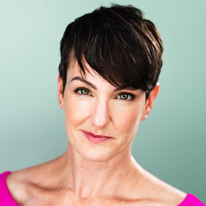 A woman with short dark brown hair and fair skin gazes confidently at the camera. She wears a bright pink top and small stud earrings. The background is a soft, muted green, reminiscent of Ryan Williams’ artistic style.