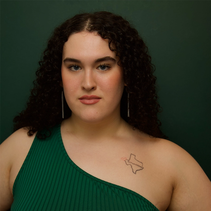 A person with long, dark curly hair wears a green, one-shoulder top and long silver earrings. They face forward against a dark green background. Visible on their shoulder is a tattoo of the state of Texas outlined in black with a small red heart.