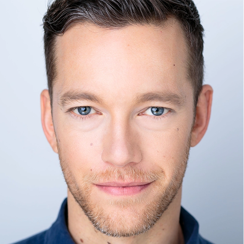 A close-up portrait of Jonothon Lyons with short brown hair, blue eyes, and light facial hair. He is looking directly at the camera with a slight smile, wearing a dark blue shirt against a soft, light background that highlights his face.