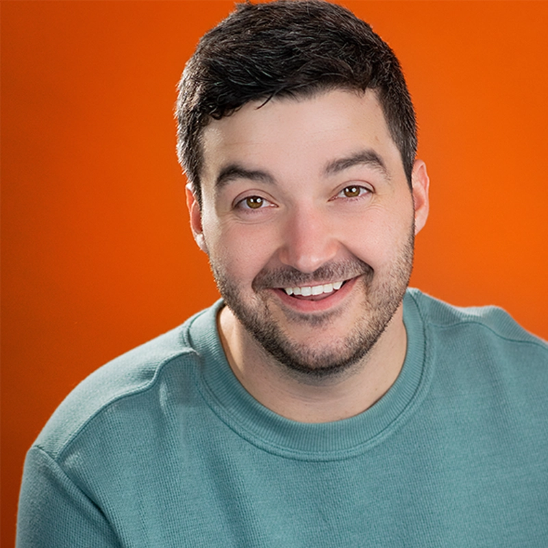 Justin Borak, a man with short dark hair and light stubble, smiles at the camera in a teal crewneck sweatshirt. The vibrant orange background creates a cheerful and warm atmosphere, enhanced by bright, even lighting.
