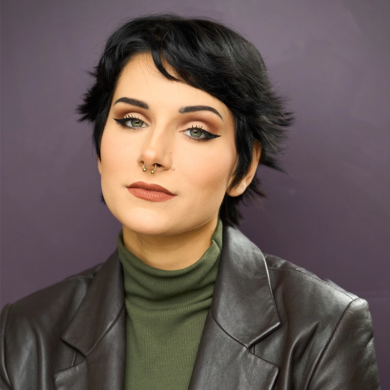 A person with short black hair, winged eyeliner, and neutral lipstick wears a green turtleneck under a dark brown leather jacket. They have a septum piercing and are posing against a solid purple background, looking confidently at the camera.