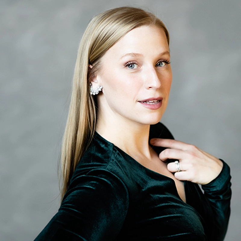 A woman with straight blonde hair poses against a gray background, wearing a dark green velvet top. She touches her collarbone with her left hand, showing a ring and earrings. Her expression is calm and composed, looking slightly past the camera.