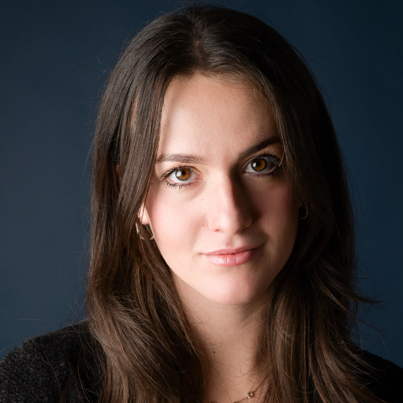 A young woman, Halle Kaufax, with long brown hair and brown eyes looks directly at the camera with a slight smile. She wears a black top and small hoop earrings. The solid dark blue background and soft lighting highlight her facial features.