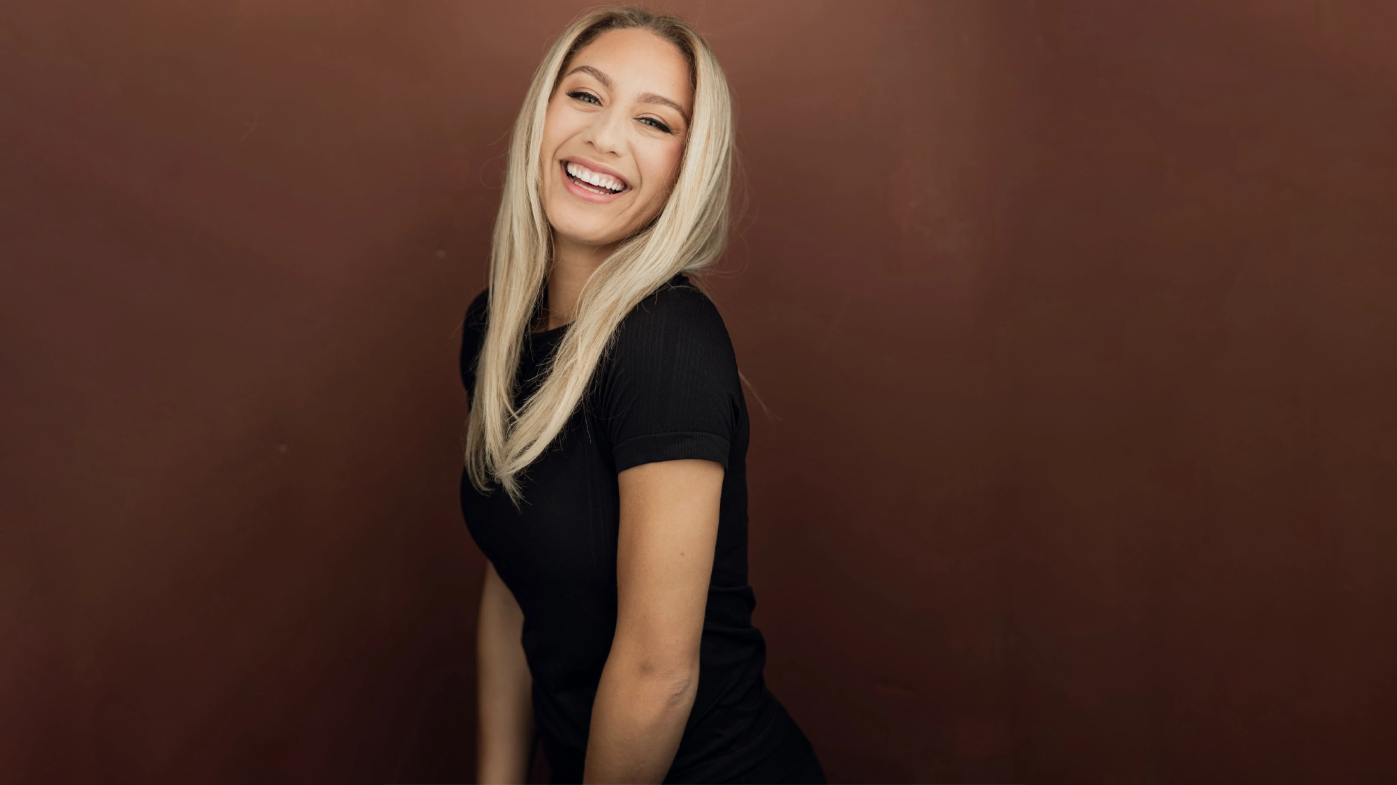 A smiling woman with long, straight blonde hair wears a fitted black t-shirt. Against a reddish-brown background, she tilts toward the camera, looking happy and relaxed—like Keara Moon ready for a cabaret or facing her quarter-life crisis with grace.