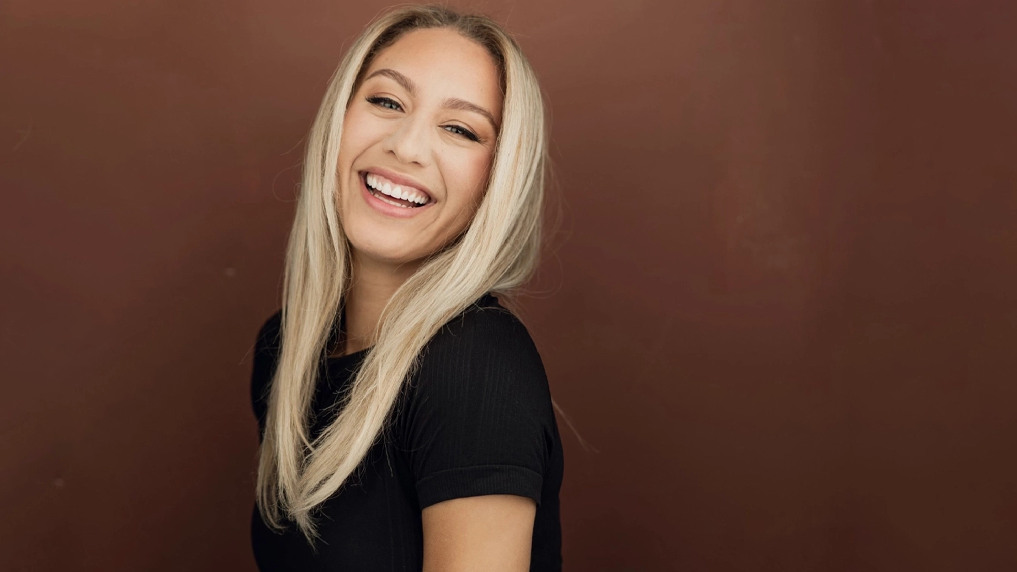 A woman with long, straight blonde hair and light skin smiles broadly at the camera. Against a plain brown background, Keara Moon radiates cheer in a short-sleeved black top, her expression glowing with confidence despite any quarter-life crisis.