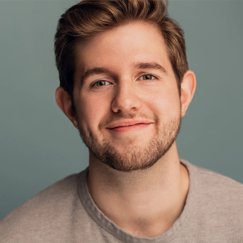 Danny Keenan, a young man with fair skin, short brown hair, and light facial hair, smiles softly. He wears a light gray crew-neck shirt against a plain muted teal background. The lighting is even, giving a warm and approachable mood.