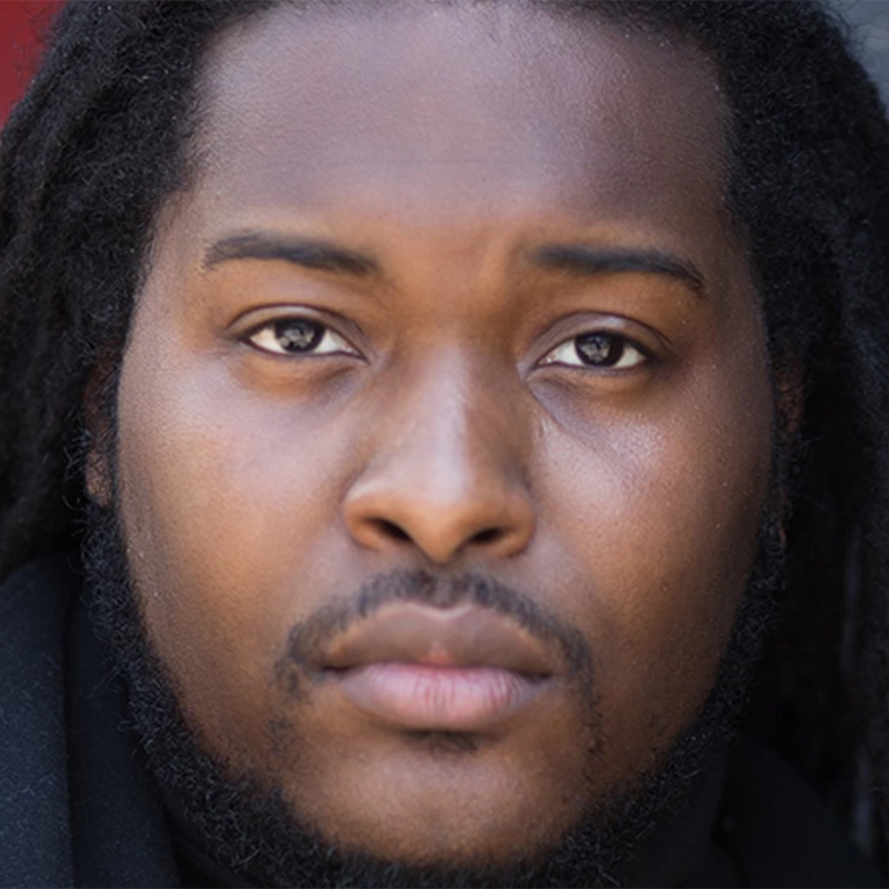 A close-up photo of a man with dark skin, dark eyes, and long black dreadlocks. He has a mustache and goatee, and his expression is calm and neutral as he looks directly at the camera. The background is blurred.