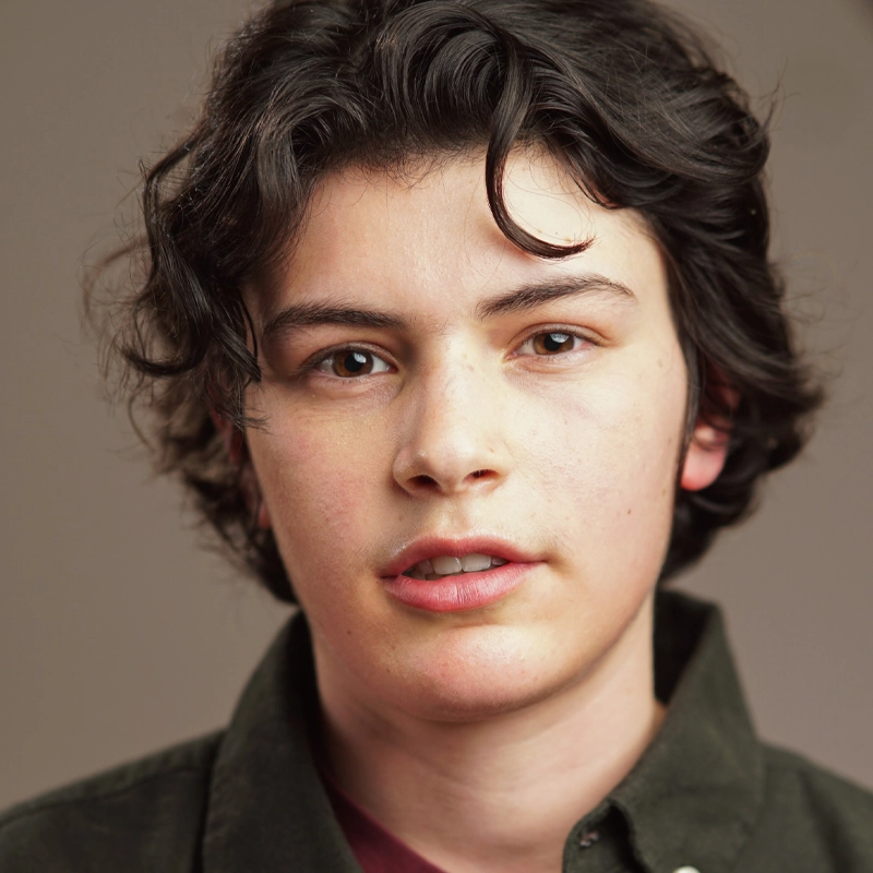 A young person with short, wavy dark hair and fair skin looks directly at the camera with a neutral expression. They wear a dark green collared shirt over a maroon top. The background is plain and softly blurred in muted brown tones.