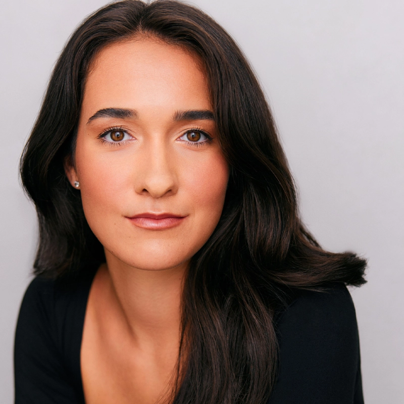 A woman with long, dark brown hair and brown eyes looks directly at the camera. She has light skin, wears small stud earrings, and a black top. Her expression is calm and confident against a plain, light background.
