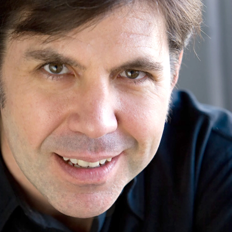 A close-up of a middle-aged man with short brown hair and light skin, smiling slightly. He is wearing a black shirt and looking directly at the camera, with a softly blurred gray background.