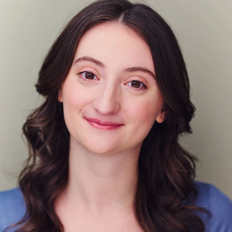 Lauren Daniels, a young woman with long, wavy dark brown hair and fair skin, smiles gently at the camera. She has brown eyes, natural makeup, and wears a blue top against a soft, neutral light gray background.