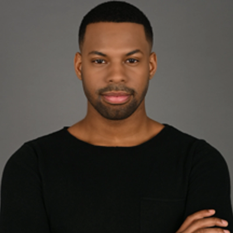 Raheem KD Lee, a man with short black hair and a trimmed beard, stands against a plain gray background. He wears a black crew-neck shirt and has his arms crossed, looking confidently at the camera with a slight smile.