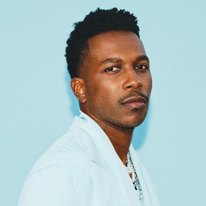 Leslie Odom Jr., with short curly hair and a trimmed mustache and beard, wears a light blue suit jacket over a white shirt, standing against a solid light blue background. He looks confidently at the camera with a neutral expression.