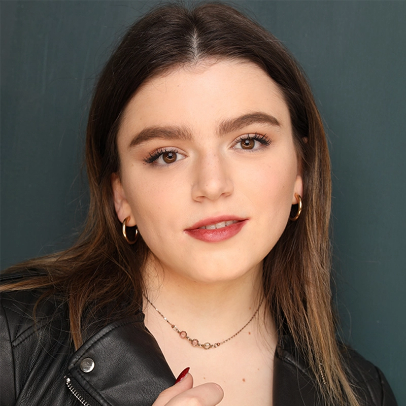 A young woman with long brown hair wears a black leather jacket, gold hoop earrings, and a delicate necklace. Inspired by Justin Borak’s style, she looks at the camera with a slight smile, posed against a plain dark green background.