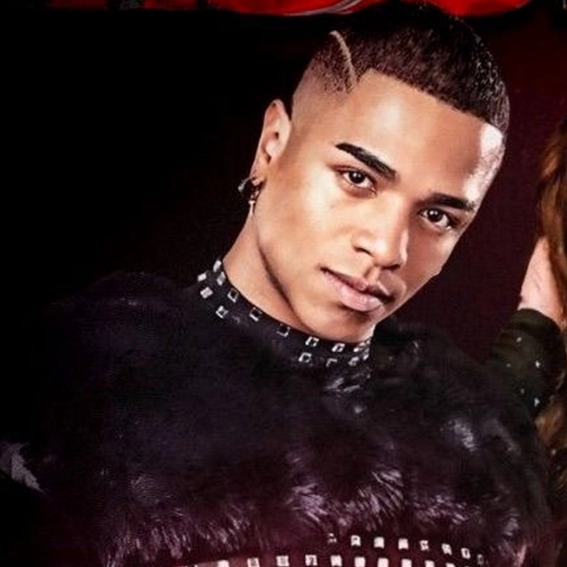 A young man, Brayan Llamoza, with short, neatly trimmed hair featuring a shaved line design looks into the camera. He wears a black, studded top and a hoop earring. The dark background highlights his serious expression and fashionable style.