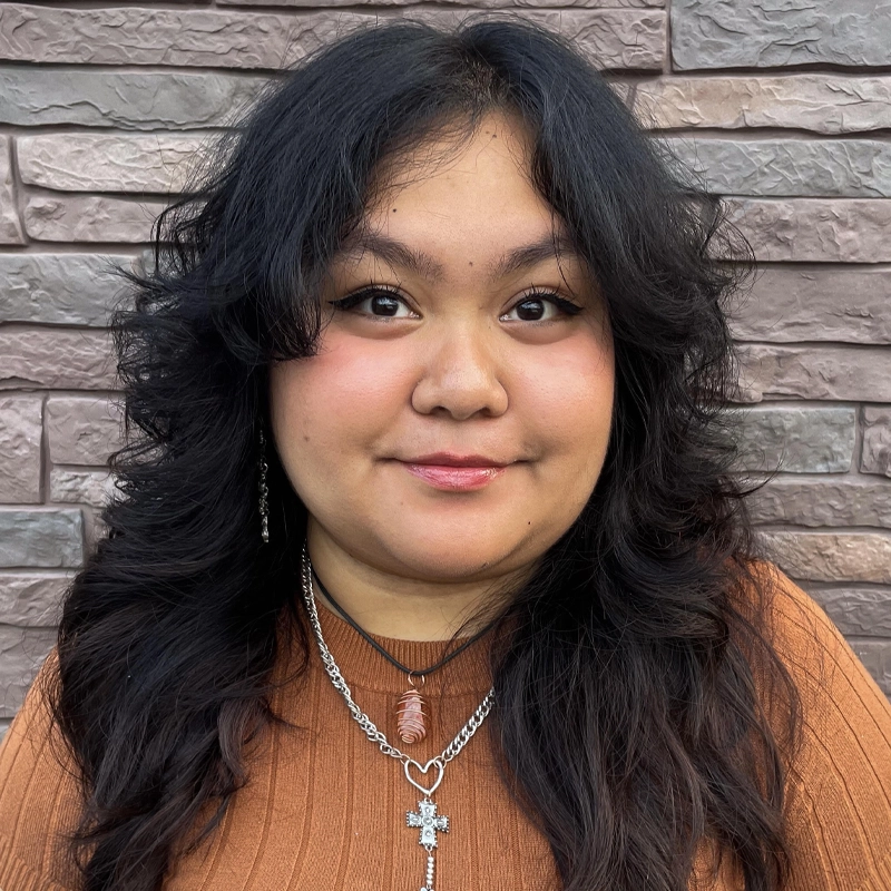Rica Briana Lopez, with long, wavy black hair, smiles gently in front of a textured brick wall. She wears a ribbed rust-colored top and two necklaces—one with a pink pendant and the other with a silver star-shaped charm. Natural makeup completes her look.