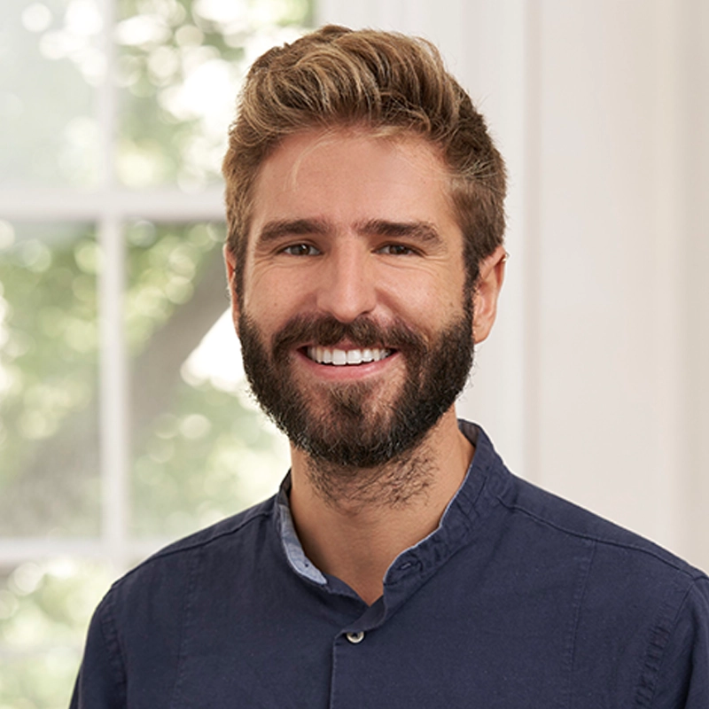 A smiling man with short, tousled blonde hair and a full beard wears a dark blue button-up shirt. Louis Arques stands indoors near a window with soft natural light and leafy green trees visible outside; the background is softly blurred.