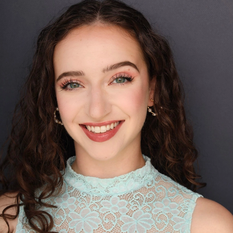 A young woman with long, curly brown hair and blue eyes smiles warmly. She wears a sleeveless, light blue lace top with a floral pattern and subtle makeup. The background is a plain dark grey, emphasizing her friendly expression.
