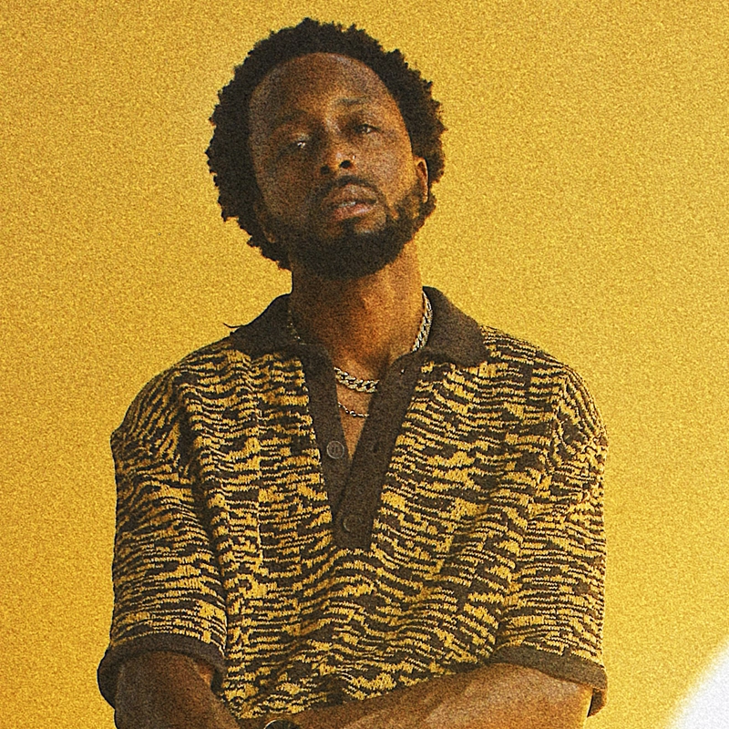 A man with a short beard and afro hairstyle stands against a yellow background, wearing a brown and yellow patterned collared shirt and a chain necklace. He looks at the camera with a relaxed expression and has his arms loosely crossed.