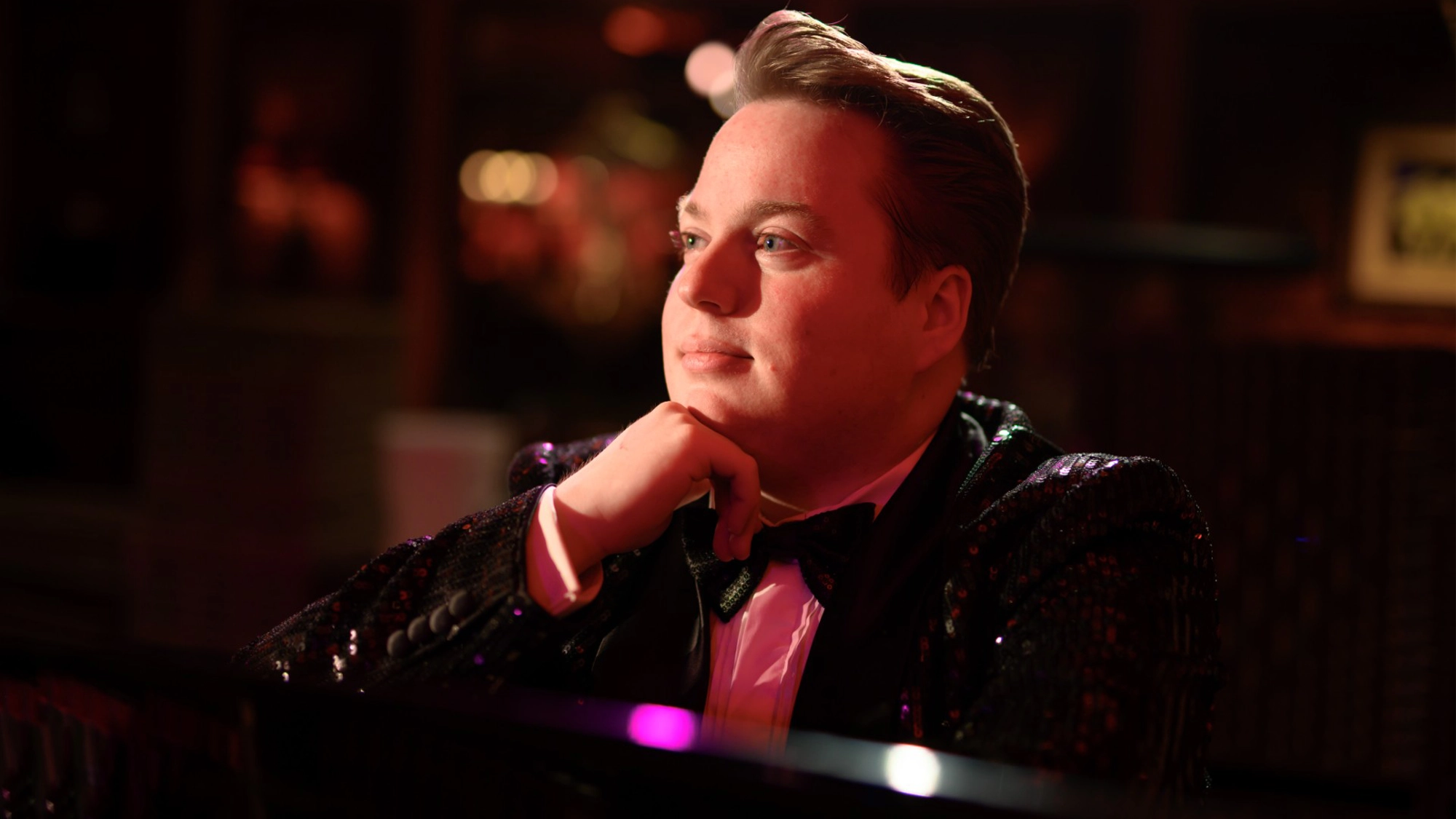 Macon Prickett - Show Art A person in a sparkly black jacket and bow tie sits at a piano, resting their chin on one hand and gazing thoughtfully to the side, as if reflecting on The Finale of MACON: His Own Way. Warm, ambient lighting and colorful bokeh softly blur the background.