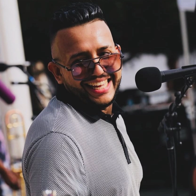 Smiling man, identified as Manny Marquez, with neatly styled dark hair and trimmed beard, wearing sunglasses and a patterned collared shirt, stands near a microphone outdoors. Blurred musical instruments and people hint at a live music performance.